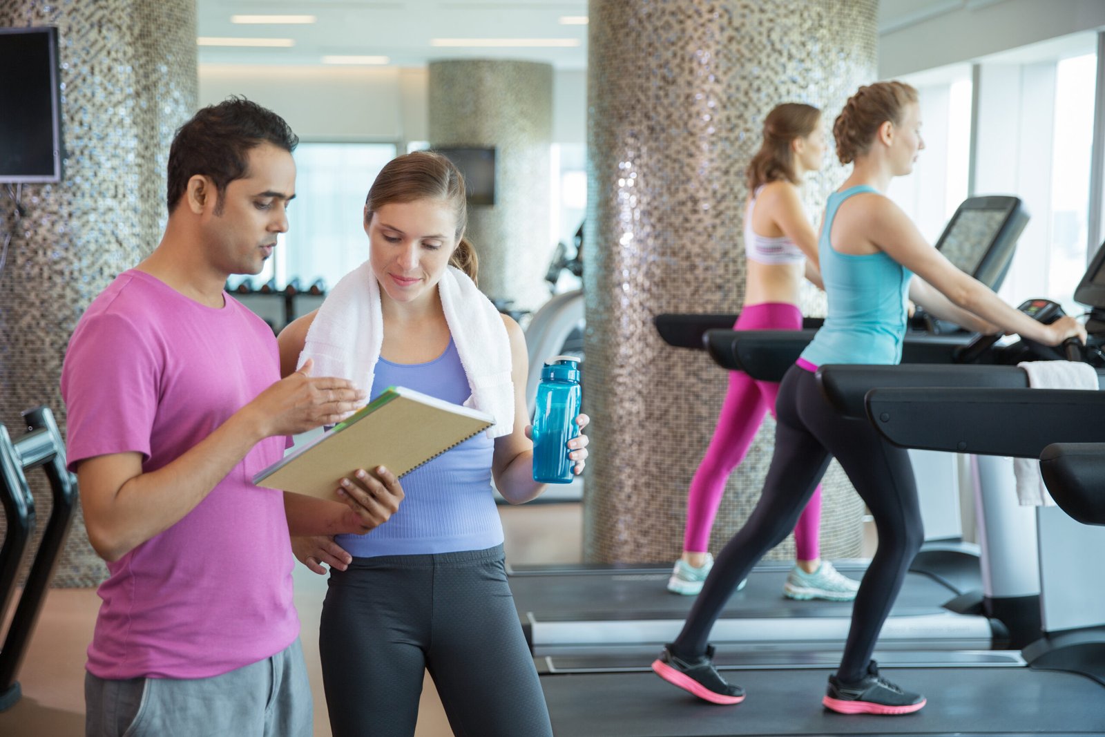 Sporty young Caucasian woman with towel talking to her male trainer in gym. He showing results of her workout. Women running on treadmills in background